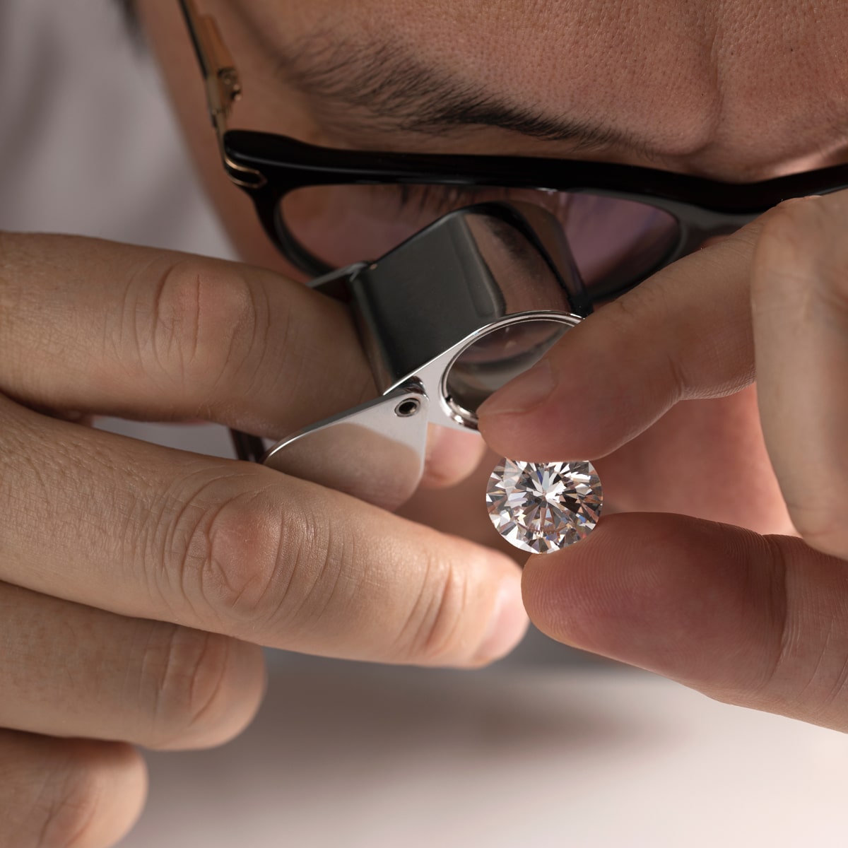 © Juwelier Wenthe Eine Brillenträgerin oder ein Brillenträger betrachtet einen runden, geschliffenen Diamanten mit der Lupe eines Juweliers und hält den Diamanten vorsichtig zwischen den Fingern.