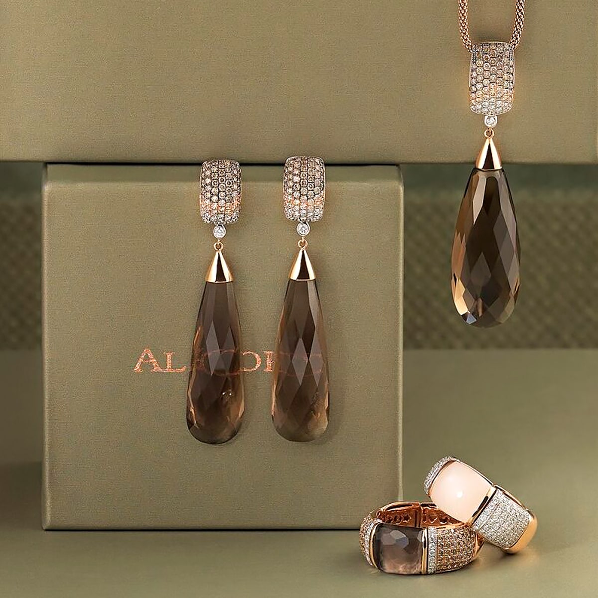2025 © Al Coro Elegantes Schmuckset von Al Coro mit tropfenförmigen Ohrringen aus braunen Edelsteinen, einem passenden Anhänger an einer Kette und zwei Ringen mit großen braunen und blassrosa Steinen, alle mit funkelnden Kristallen verziert, in einer grünen Schachtel.