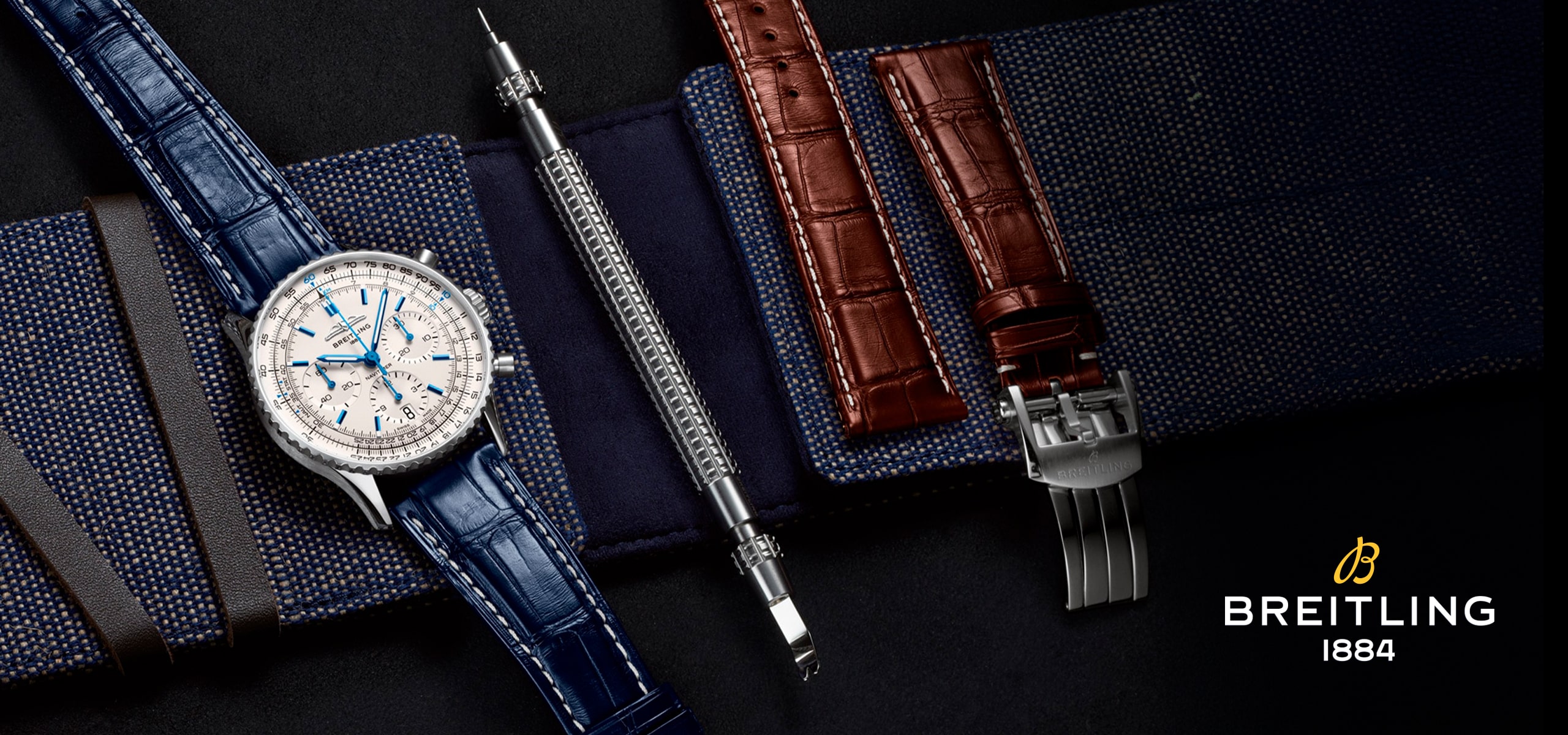 2025 © BREITLING Eine Breitling-Uhr mit blauem Lederarmband wird neben einem Werkzeug zum Wechseln des Armbands, einem braunen Lederarmband und einer Schließe gezeigt, die alle auf einem strukturierten marineblauen Stoff liegen. Das ikonische Breitling 1884 Logo erscheint in der unteren rechten Ecke.