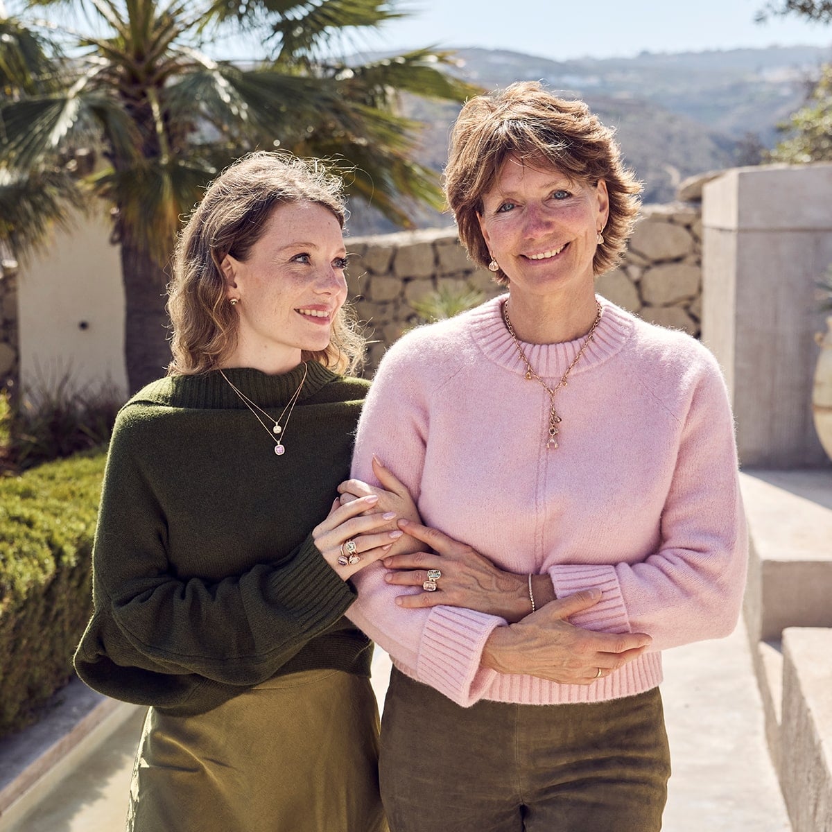 2025 © CAPOLAVORO Zwei Frauen stehen Arm in Arm im Freien und lächeln in die Sonne. Die eine trägt einen rosa Pullover, die andere einen grünen - ein Capolavoro-Moment vor Palmen, Steinmauern und sanften Hügeln im Hintergrund.