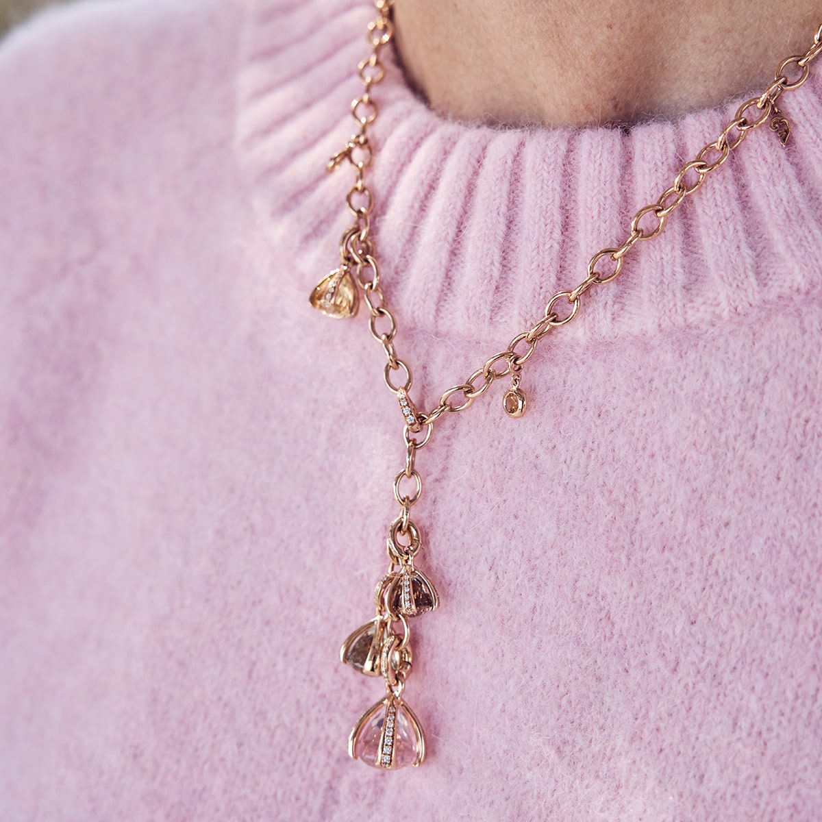 2025 © CAPOLAVORO Nahaufnahme einer Person, die eine Capolavoro-Goldkette mit mehreren baumelnden Anhängern trägt, gepaart mit einem pastellrosa Strickpullover.