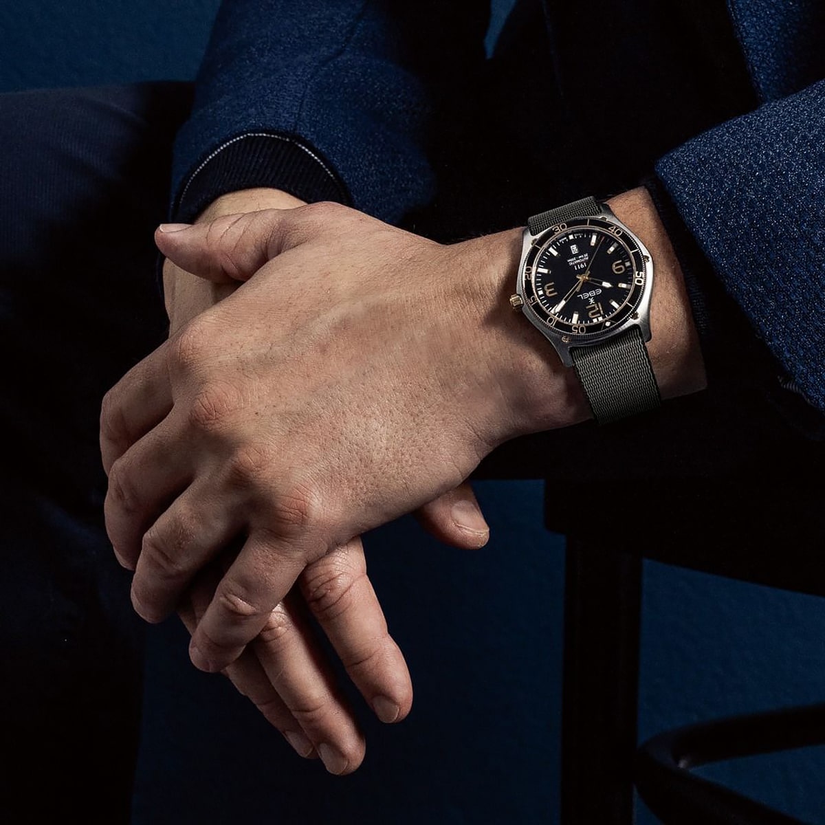 2025 © EBEL Eine Person in einem dunkelblauen Anzug sitzt mit gekreuzten Händen da und zeigt eine elegante schwarze Ebel-Armbanduhr mit dunklem Armband und silbernem Gehäuse an ihrem linken Handgelenk.