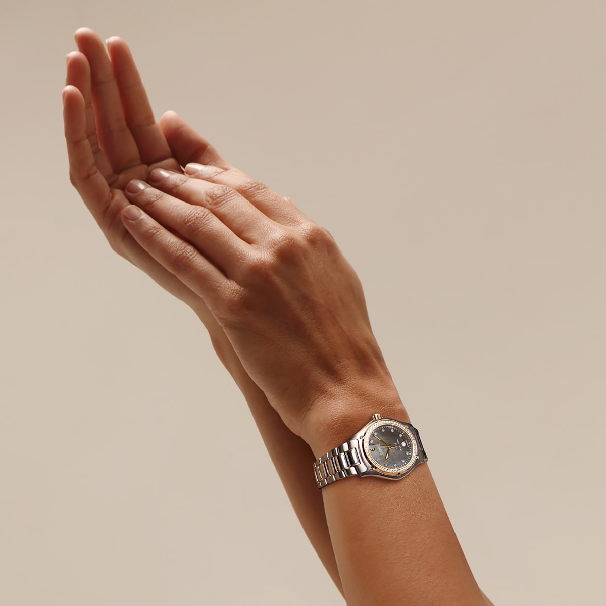 2025 © EBEL Nahaufnahme von zwei Händen, die sich vor einem schlichten Hintergrund zusammenhalten, an einem Handgelenk eine elegante Ebel-Armbanduhr in Silber und Gold mit einem runden schwarzen Zifferblatt.