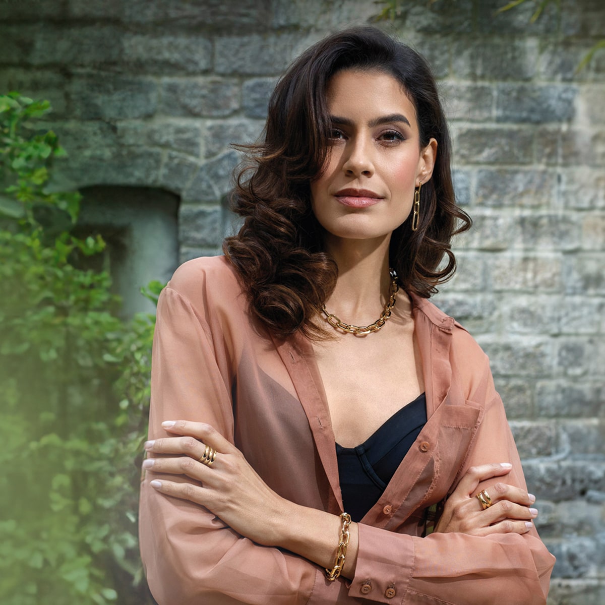 2025 © Elaine Firenze Elaine Firenze, mit gewelltem braunem Haar, Goldschmuck und einer durchsichtigen pfirsichfarbenen Bluse über einem schwarzen Top, steht selbstbewusst im Freien vor einer Steinmauer und Grünpflanzen, die Arme verschränkt, während sie in die Kamera blickt.