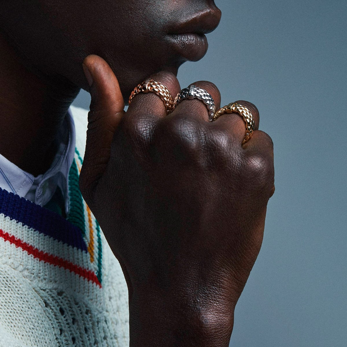 2025 © FOPE Eine Person mit dunkler Hautfarbe stützt ihr Kinn auf ihre Hand und zeigt mehrere goldene und silberne Ringe mit Fope-Struktur. Sie trägt einen weißen Strickpullover mit blauen, roten und gelben Streifen über einem Kragenhemd.