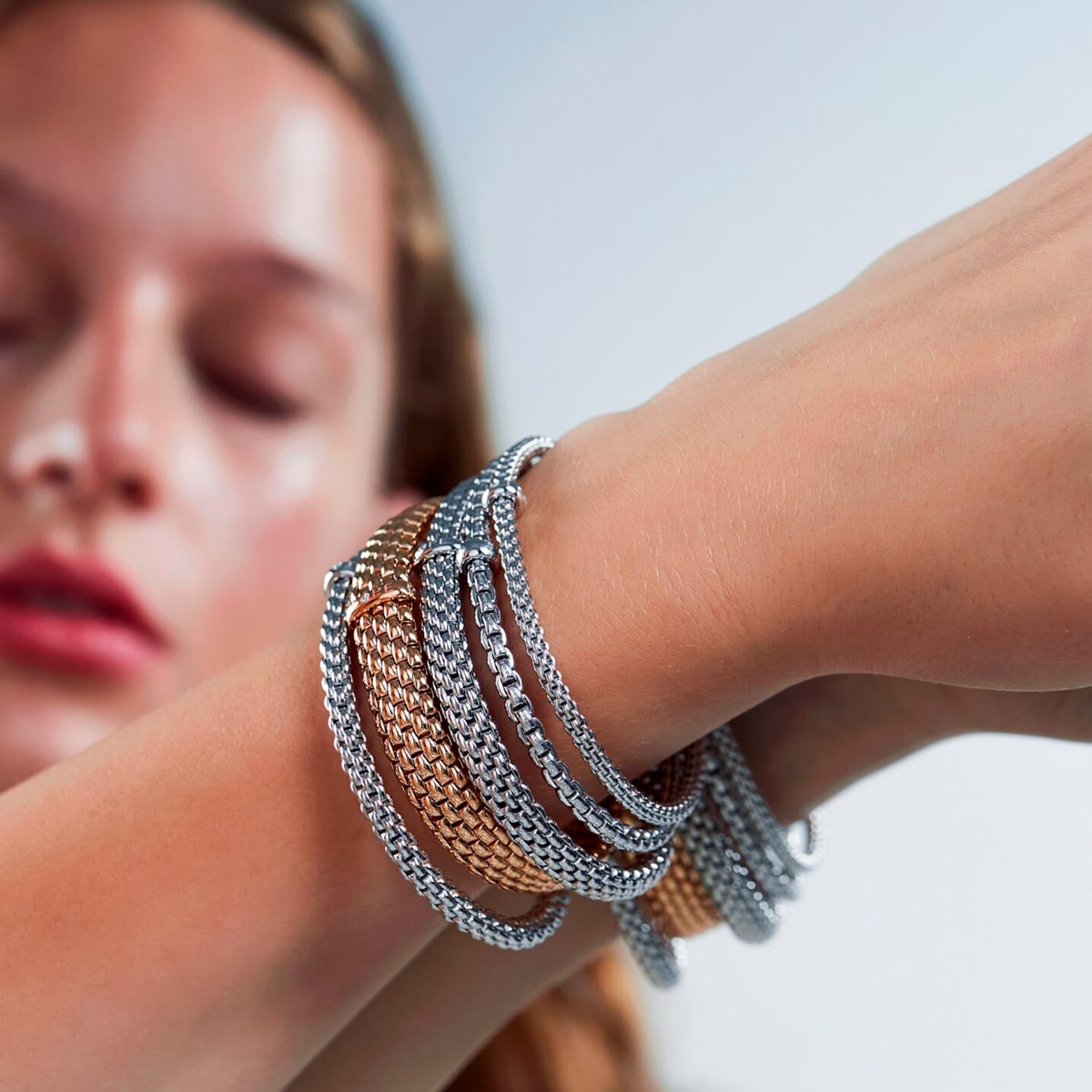 2025 © FOPE Nahaufnahme des Handgelenks einer Frau, das mit mehreren goldenen und silbernen Fope-Kettenarmbändern geschmückt ist. Ihr Gesicht ist im Hintergrund elegant unscharf.