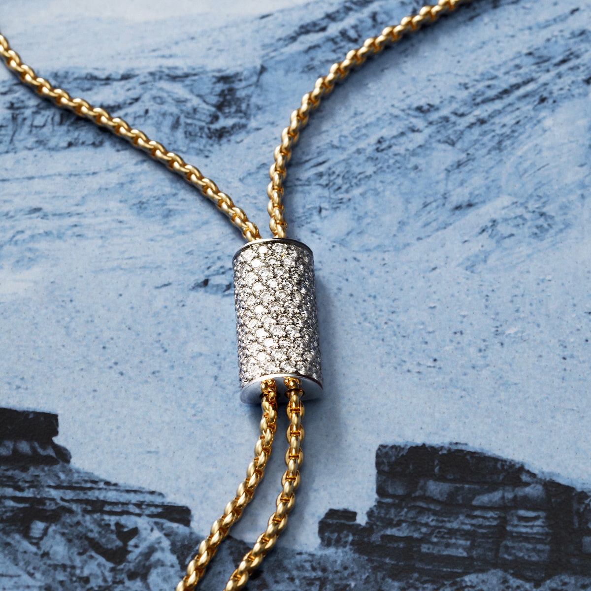 2025 © FOPE Eine goldene Fope-Kette mit einem zylindrischen, diamantbesetzten Anhänger ist auf einem blau-weißen Hintergrund mit Steinstruktur zu sehen.