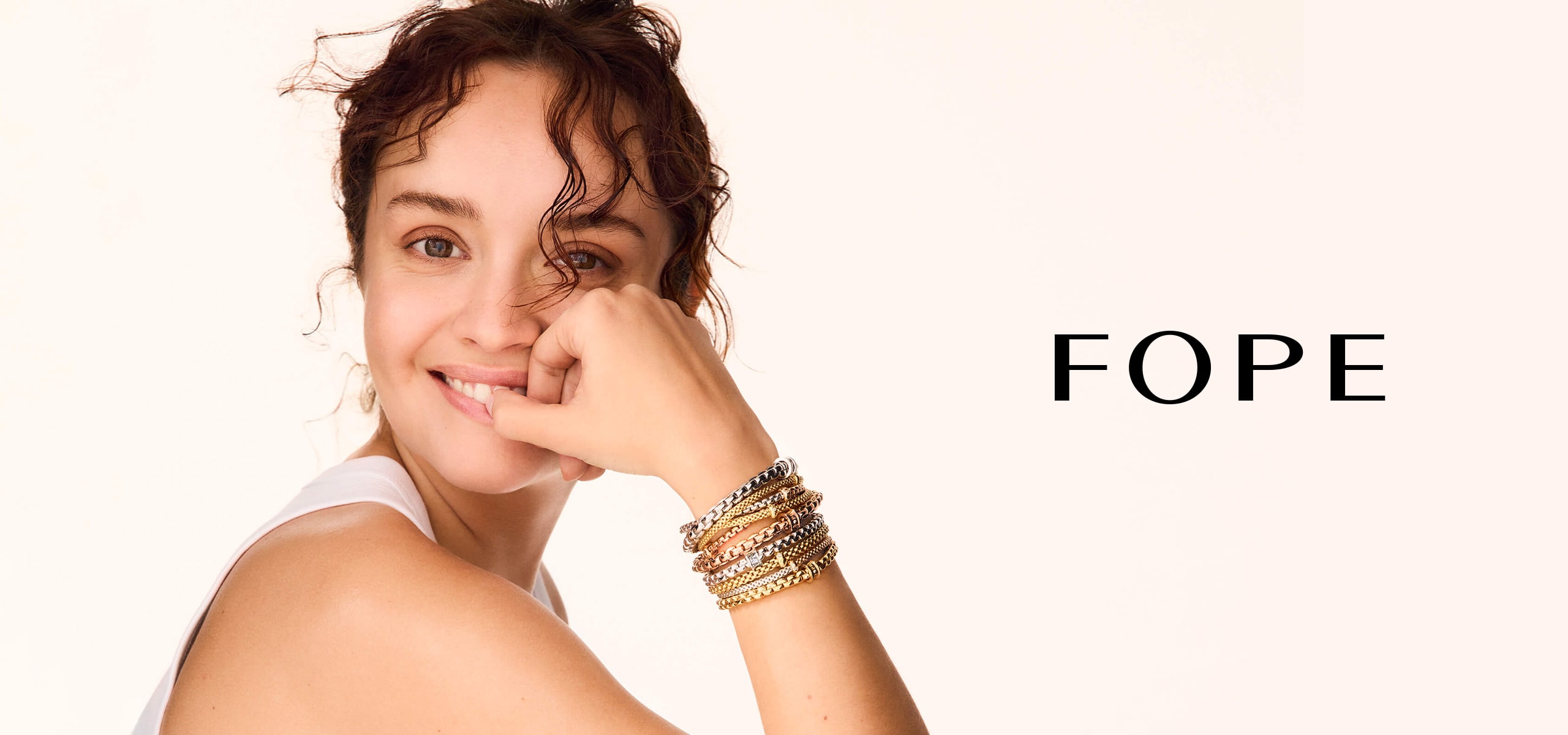 2025 © FOPE Eine Frau mit lockigem Haar stützt lächelnd ihr Kinn auf die Hand und präsentiert mehrere goldene und silberne Fope-Armbänder an ihrem Handgelenk. Neben ihr erscheint das Wort "FOPE" auf einem schlichten hellen Hintergrund.