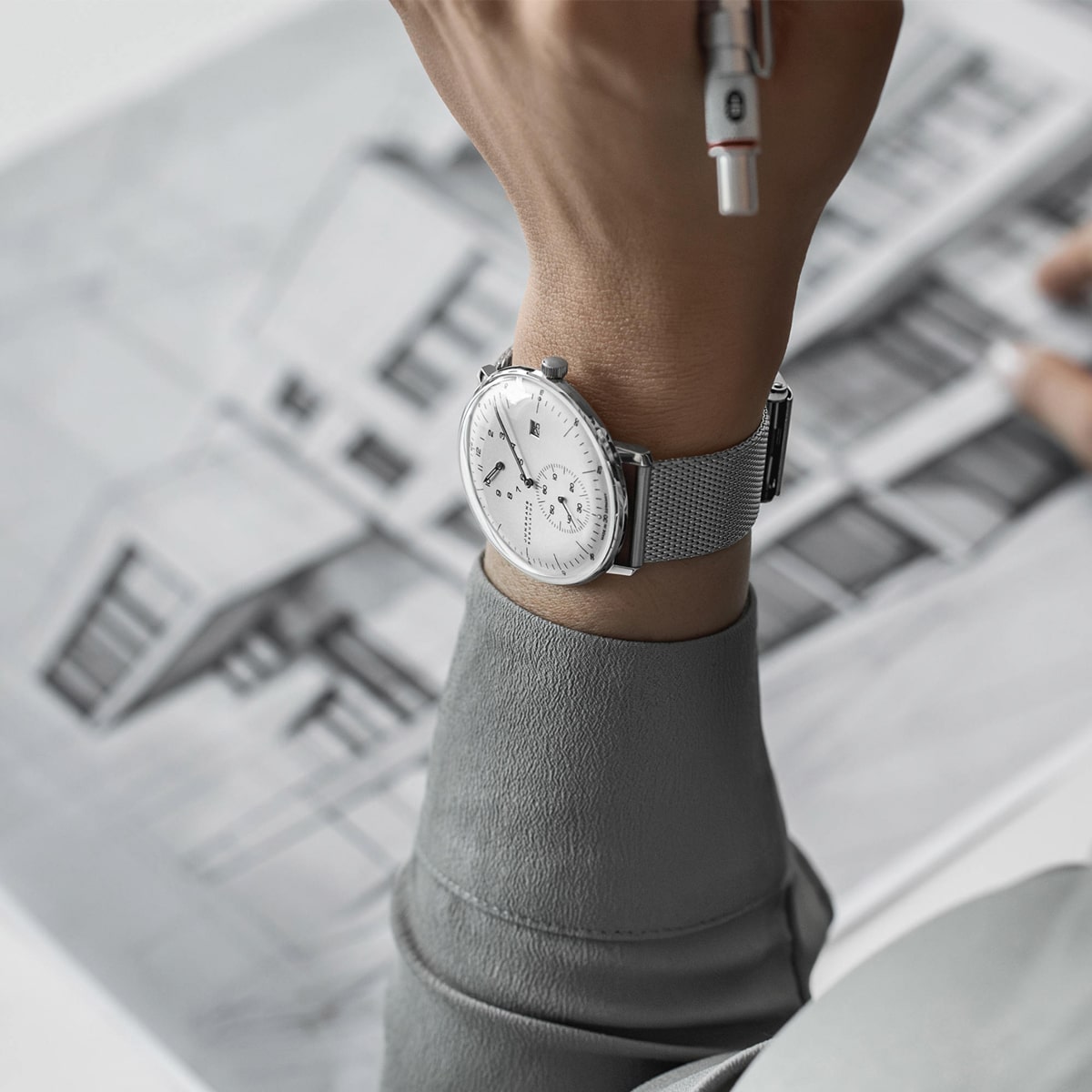 2025 © Junghans Eine Person mit einer silbernen Junghans-Armbanduhr hält einen Stift über einer architektonischen Zeichnung eines modernen Gebäudes, bekleidet mit einem hellgrauen langärmeligen Hemd.