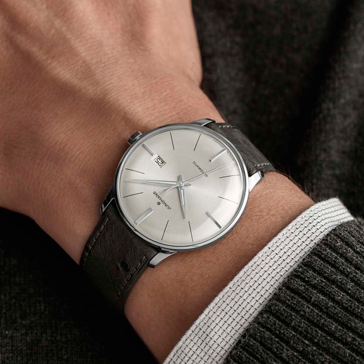 2025 © Junghans Nahaufnahme eines Handgelenks, das eine minimalistische Junghans-Armbanduhr mit silbernem Zifferblatt, schwarzem Lederarmband, silbernen Indexen und einem kleinen Datumsfenster trägt, vor einem dunklen Pullover und einer weißen Hemdmanschette.