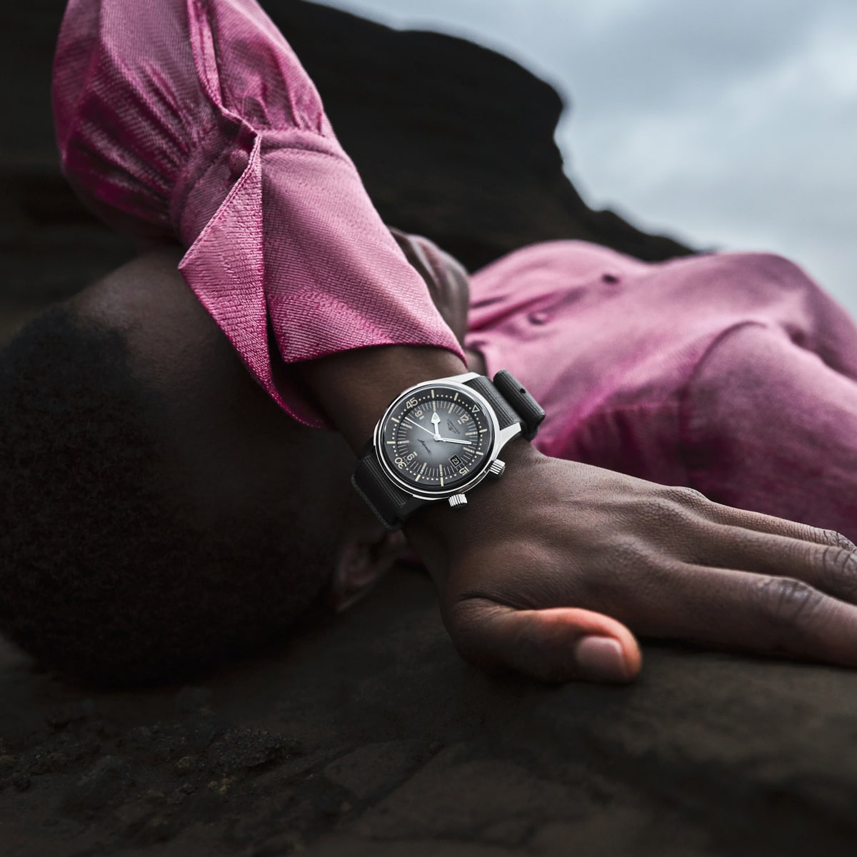 2025 © LONGINES Eine Person in einem leuchtend rosafarbenen Hemd liegt auf einem dunklen Felsen, einen Arm über dem Gesicht und präsentiert eine elegante LONGINES-Armbanduhr. Der Himmel im Hintergrund ist bewölkt und bedeckt.