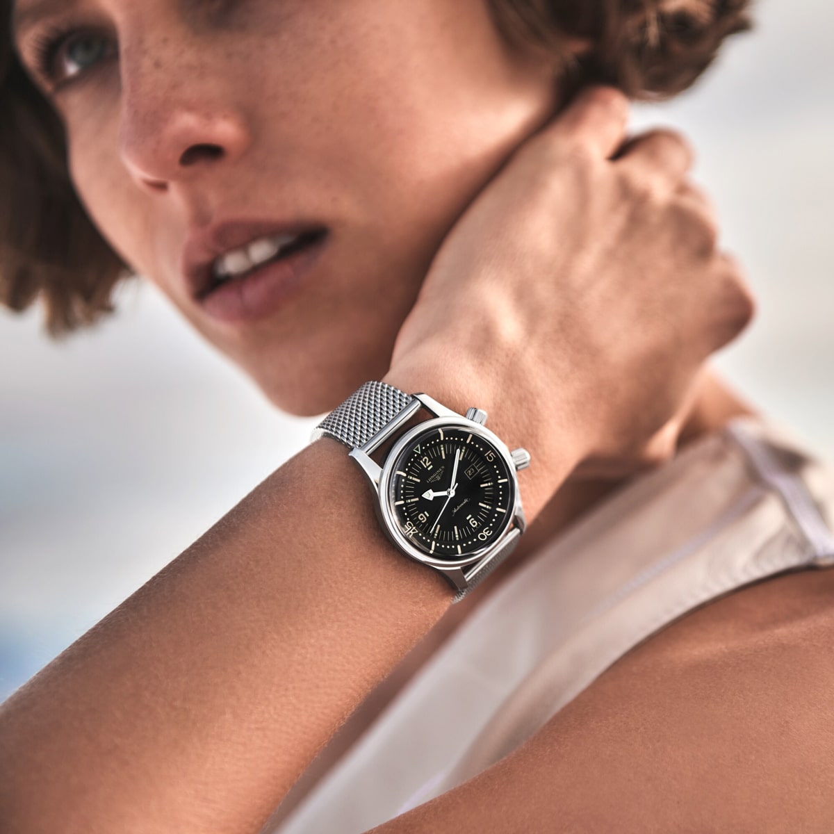 2025 © LONGINES Eine Person mit kurzen braunen Haaren trägt ein ärmelloses weißes Oberteil und eine schlichte silberne LONGINES-Armbanduhr mit schwarzem Zifferblatt, wobei sie mit der Hand im Nacken posiert. Der sanft verschwommene Hintergrund verleiht ihr eine elegante Note.