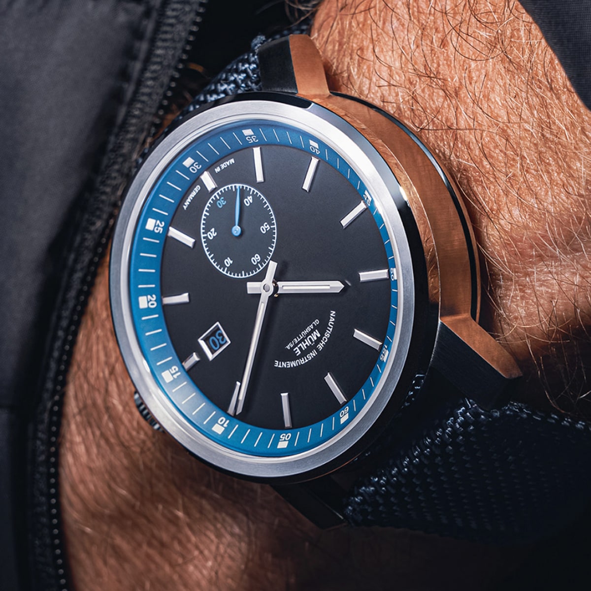 2025 © Mühle Glashütte Nahaufnahme einer luxuriösen Armbanduhr der Mühle Glashütte mit schwarzem Zifferblatt, blauen und weißen Akzenten, einem Hilfszifferblatt bei 12 Uhr und einem Datumsfenster bei 6 Uhr, getragen am Handgelenk einer Person mit einem strukturierten dunklen Armband.