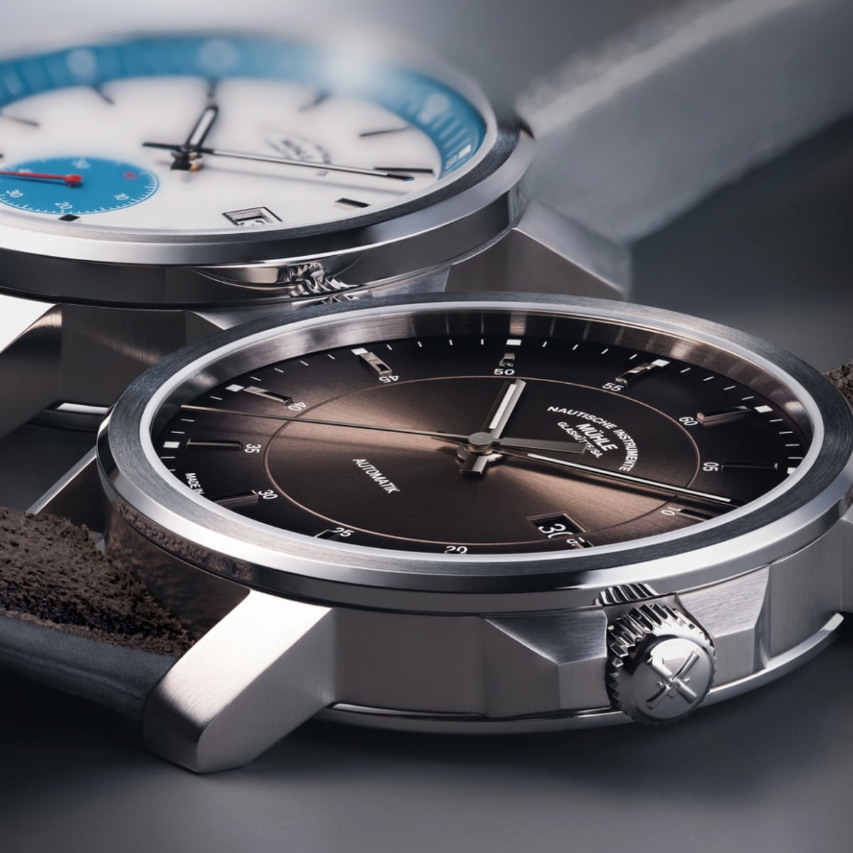 2025 © Mühle Glashütte Zwei elegante Mühle Glashütte Armbanduhren mit Metallgehäuse sind aus der Nähe zu sehen; eine hat ein braunes Zifferblatt und die andere ein weißes Zifferblatt mit blauen Akzenten. Beide zeichnen sich durch ein minimalistisches Design und sichtbare Kronenverstellungen aus.
