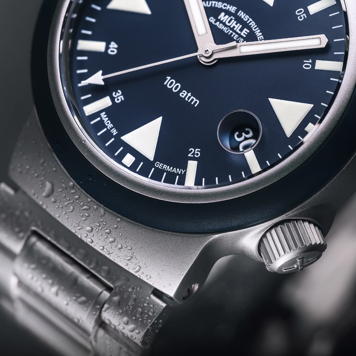 2025 © Mühle Glashütte Nahaufnahme einer silbernen Armbanduhr von Mühle Glashütte mit blauem Zifferblatt, markanten weißen Indexen und einem Datumsfenster bei 4 Uhr. Wassertropfen auf dem Metallarmband unterstreichen die beeindruckende Wasserdichtigkeit der Uhr.