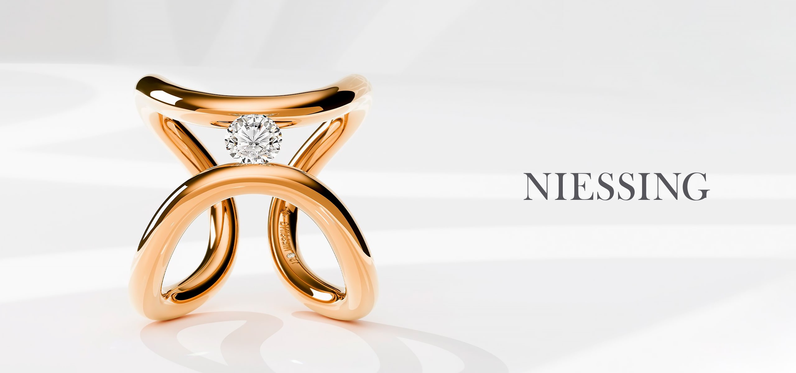 2025 © Niessing Ein goldener Ring von Niessing mit einem einzigartigen, skulpturalen Design hält einen einzelnen runden Diamanten in seiner Mitte. Rechts daneben steht in eleganten Großbuchstaben auf hellem Grund das Wort "NIESSING".