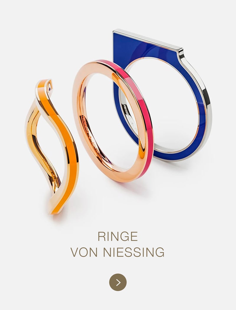 Drei moderne, geometrische Ringe von Niessing aus orangefarbenem, roségoldenem und blauem Metall sind aufrecht auf weißem Hintergrund angebracht. Der Text darunter lautet "RINGE VON NIESSING" in goldenen Großbuchstaben.