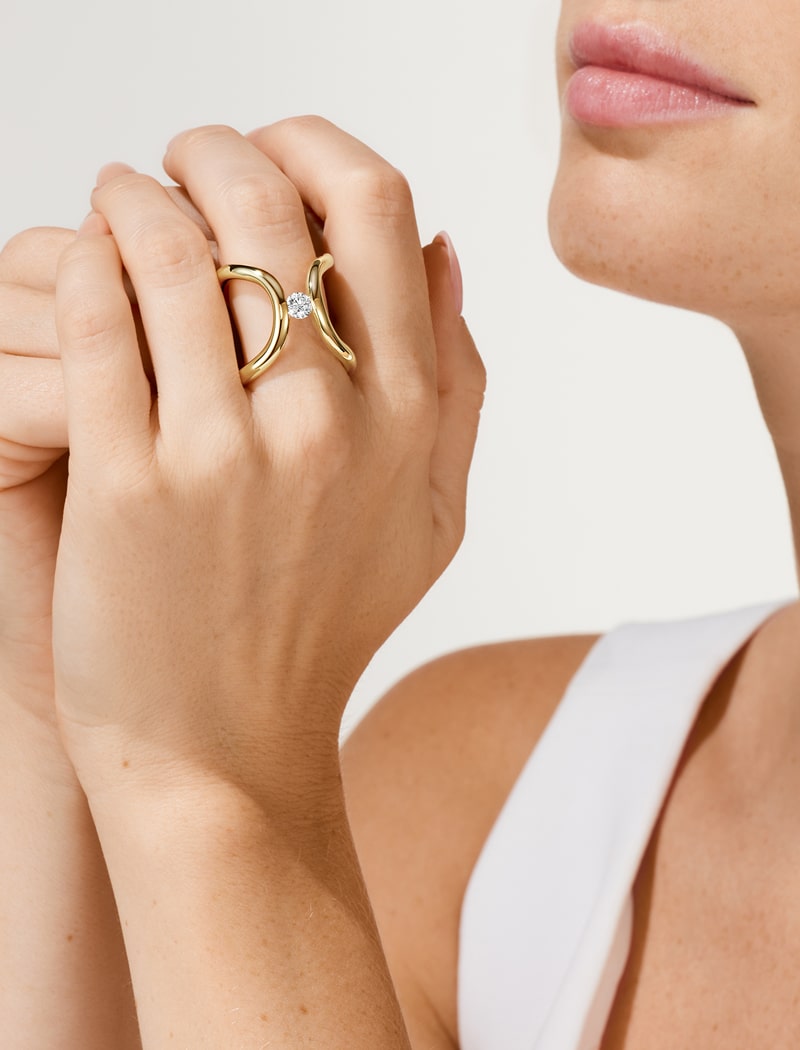 2025 © Niessing Nahaufnahme einer Frau, die ihre Hände in der Nähe ihres Gesichts zusammenhält und einen Niessing-Goldring mit einem markanten Diamanten zur Schau stellt. Sie trägt ein weißes ärmelloses Oberteil, ihre Lippen sind leicht geschürzt. Der Hintergrund ist einfarbig und hell.