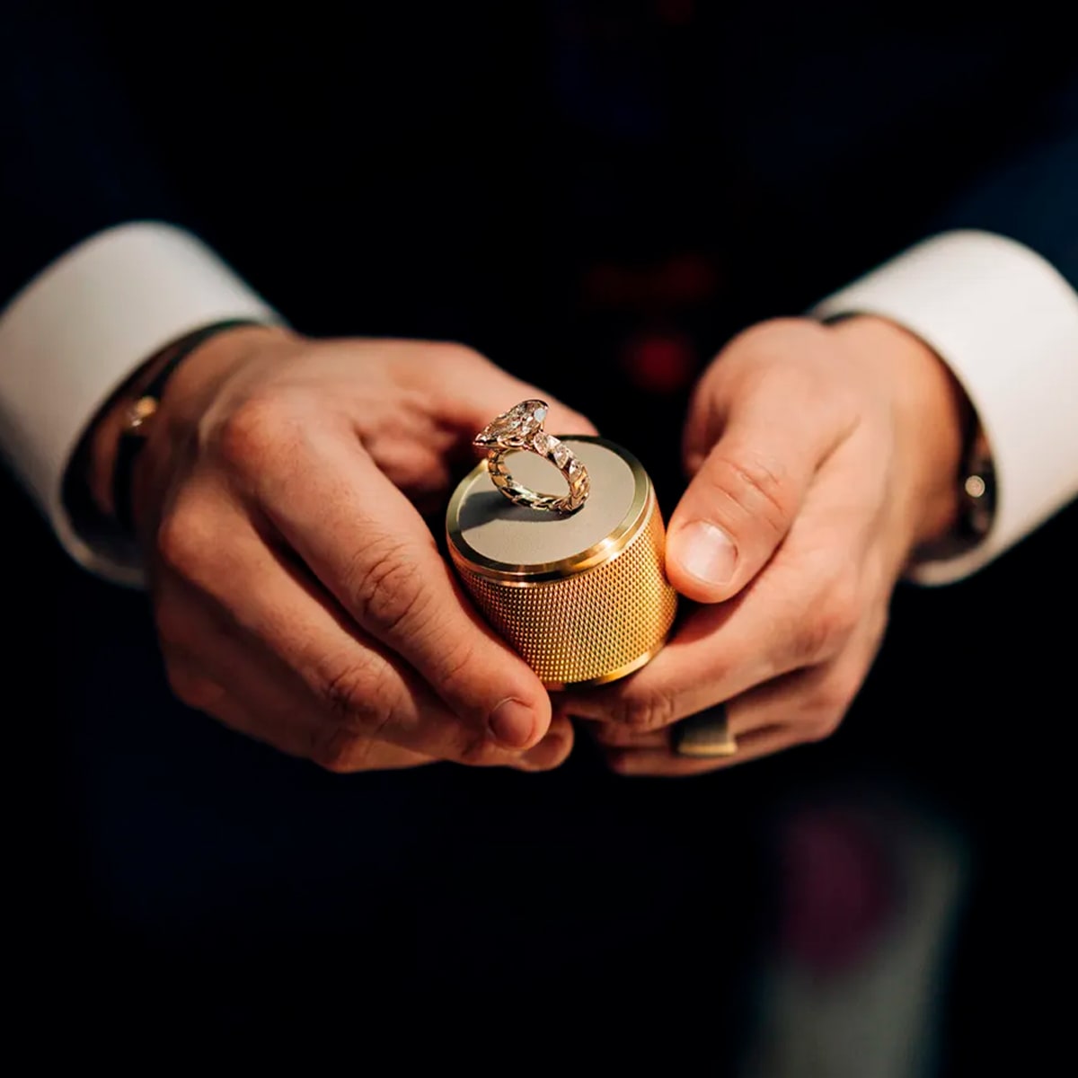 2025 © SCHAFFRATH Eine Person im Anzug hält eine goldfarbene Schaffrath-Ringschatulle in der Hand, die geöffnet ist und einen großen, funkelnden Diamantring enthüllt. Ihre Hände stehen im Mittelpunkt und heben den exquisiten Schmuck und die elegante Schachtel hervor.