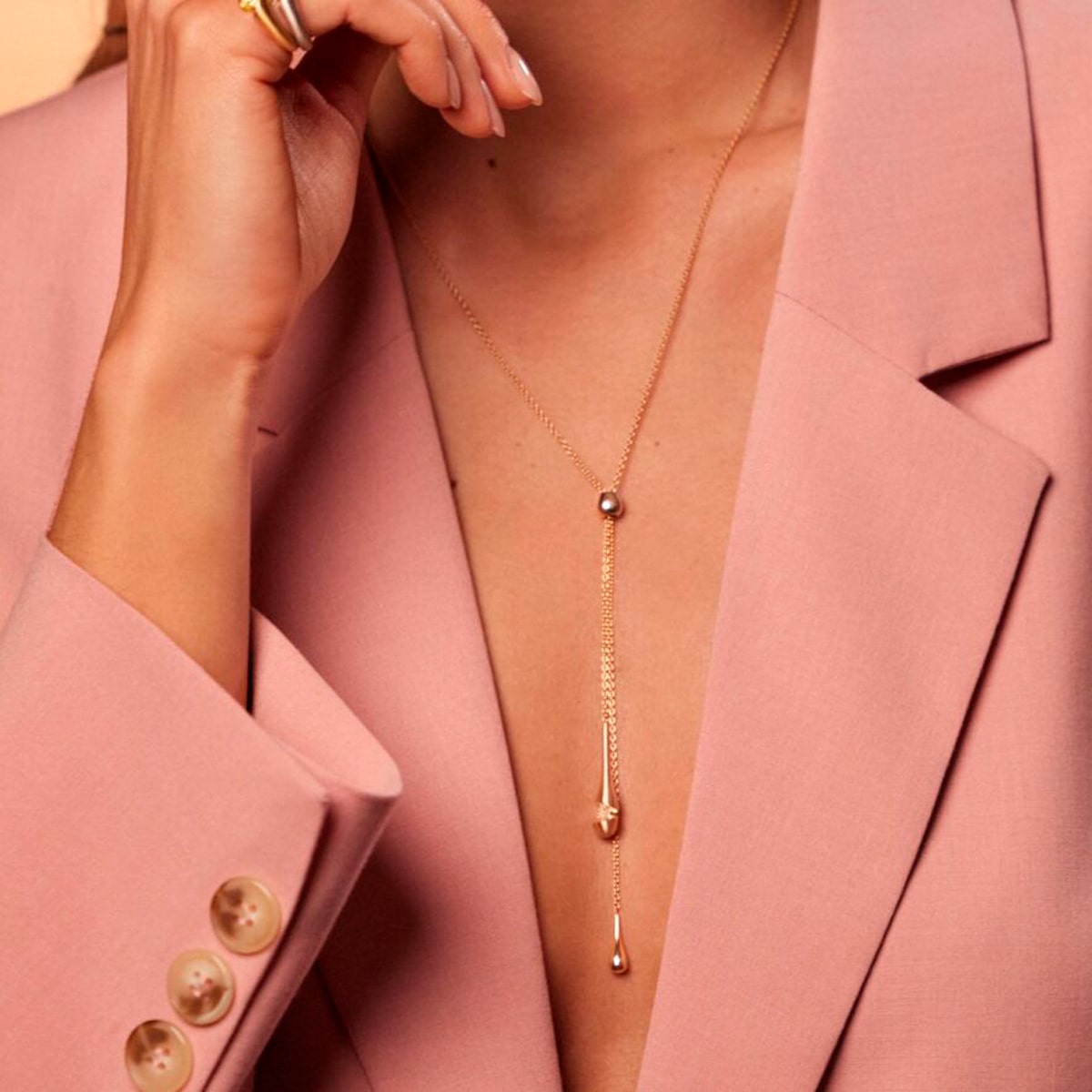 2025 © SCHAFFRATH Eine Person trägt einen rosafarbenen Blazer und eine zarte goldene Schaffrath-Lariat-Kette mit zwei baumelnden Anhängern. Die Hand ist in der Nähe des Gesichts erhoben und zeigt elegant einen goldenen Ring.
