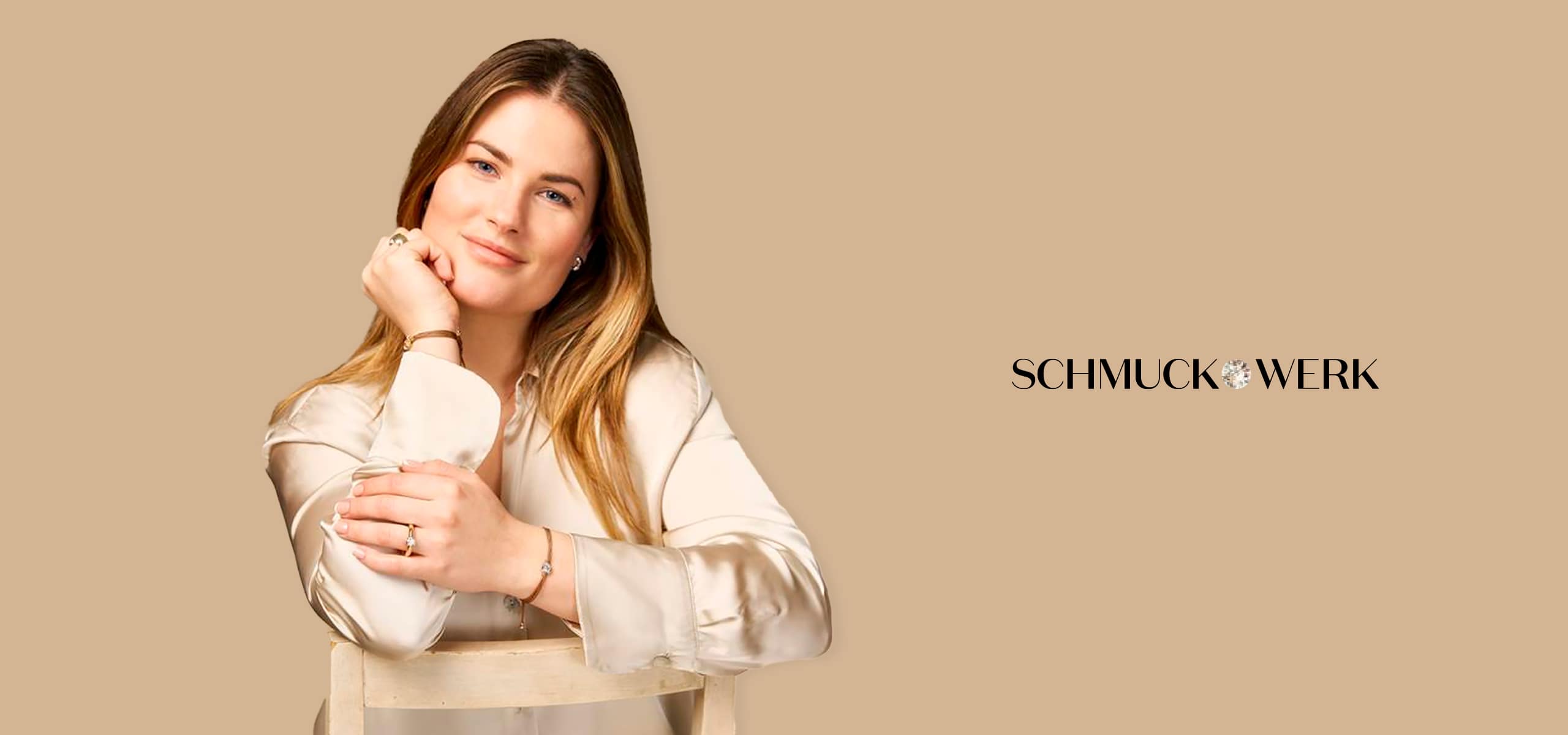 2025 © Schmuckwerk Eine Frau mit langen blonden Haaren und einer cremefarbenen Bluse lehnt sich an die Lehne eines Stuhls und lächelt sanft. Vor dem beigen Hintergrund erscheint rechts das Wort "Schmuckwerk", das eine schlichte Eleganz ausstrahlt.