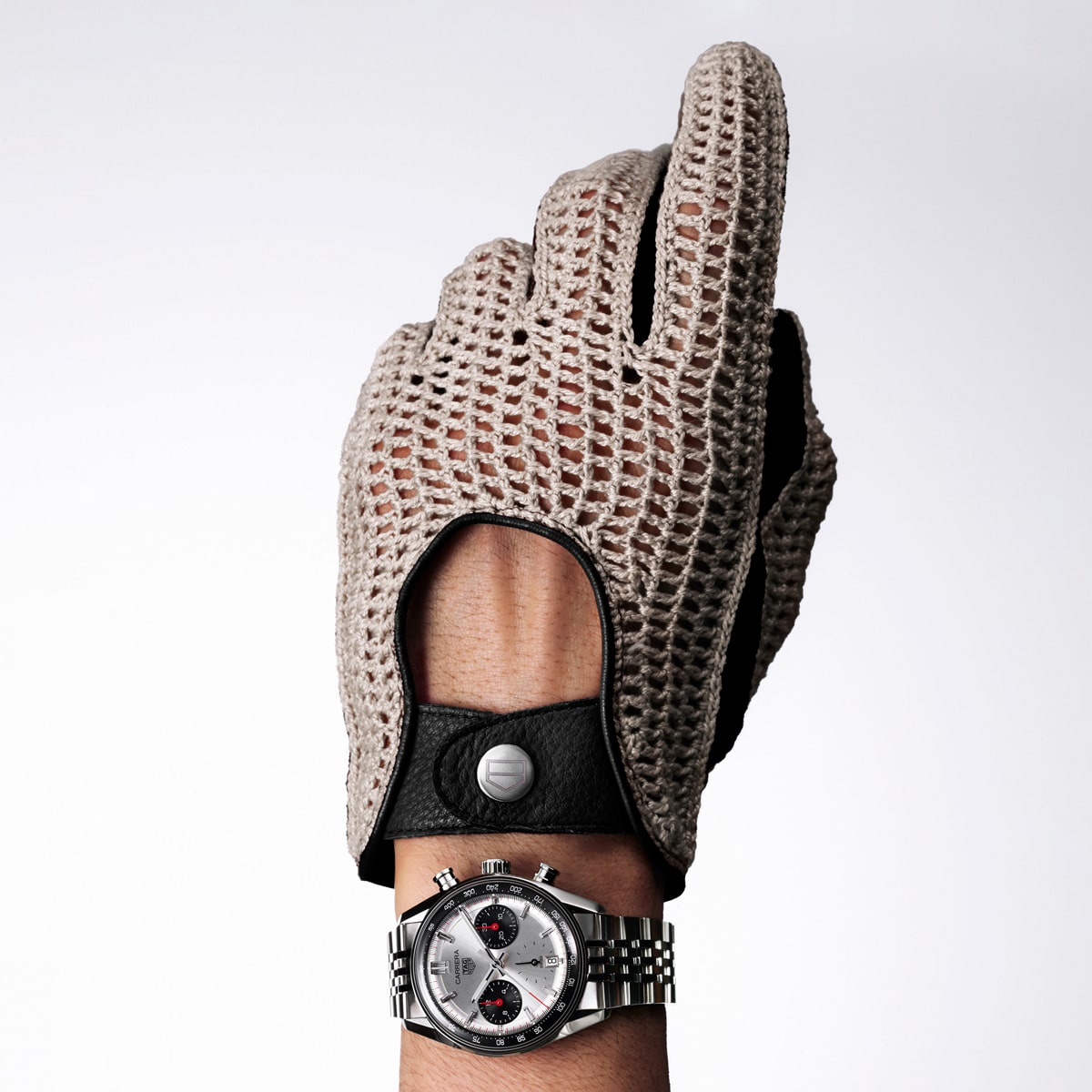 2025 © TAG HEUER Eine Hand, die einen beigen, gestrickten Fahrhandschuh mit schwarzem Lederakzent trägt, zeigt nach oben. Am Handgelenk trägt sie eine silberne Chronographenuhr mit Metallband und weißem Zifferblatt mit roten und schwarzen Ziffern.