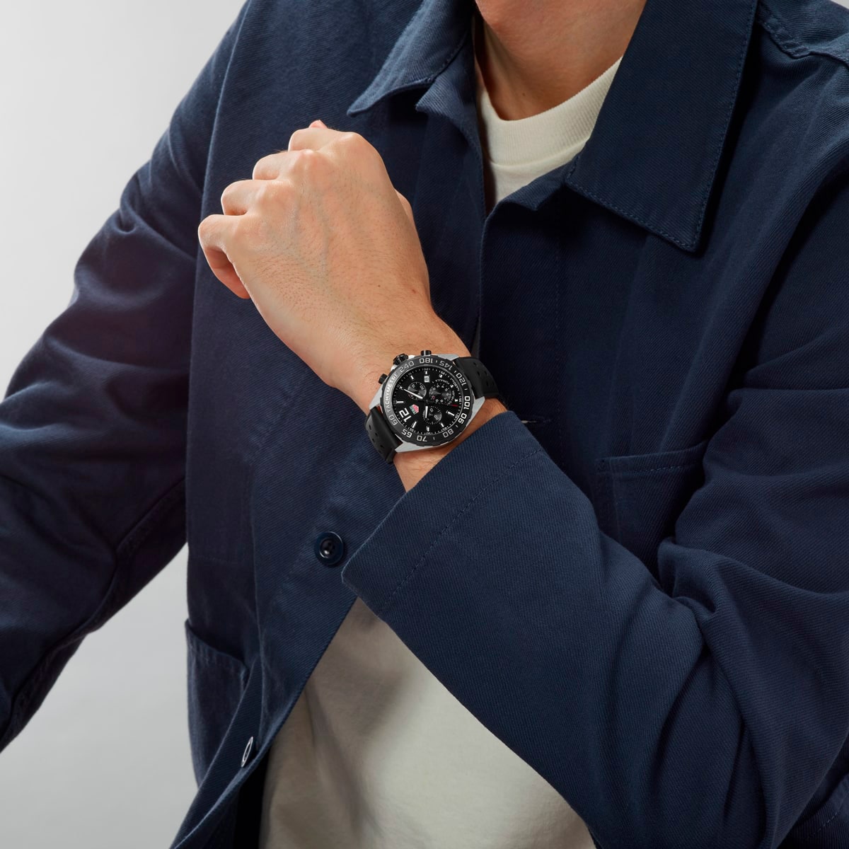 2025 © TAG HEUER Eine Person, die eine dunkelblaue Jacke und ein weißes Hemd trägt, zeigt eine schwarze Armbanduhr mit einem großen, detaillierten Zifferblatt an ihrem linken Handgelenk, wobei sie den Arm angewinkelt hält und die Hand an ihr Kinn legt.