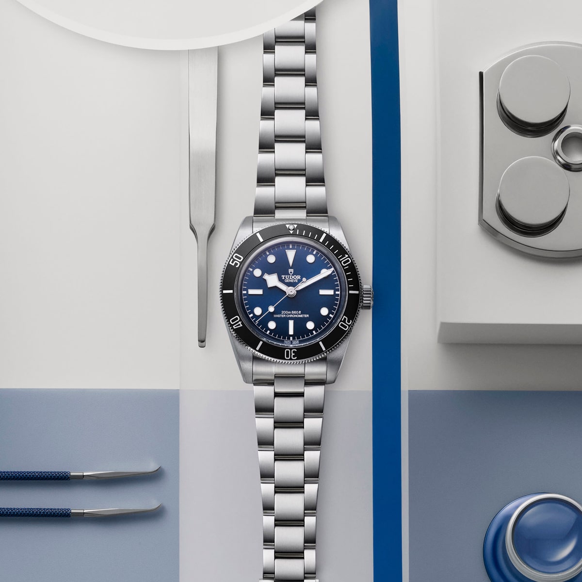 2025 © TUDOR Eine Tudor-Armbanduhr aus Edelstahl mit blauem Zifferblatt und blauer Lünette steht auf einer weiß-blauen Fläche, umgeben von Tudor-Uhrmacherwerkzeugen und -Ausrüstung.