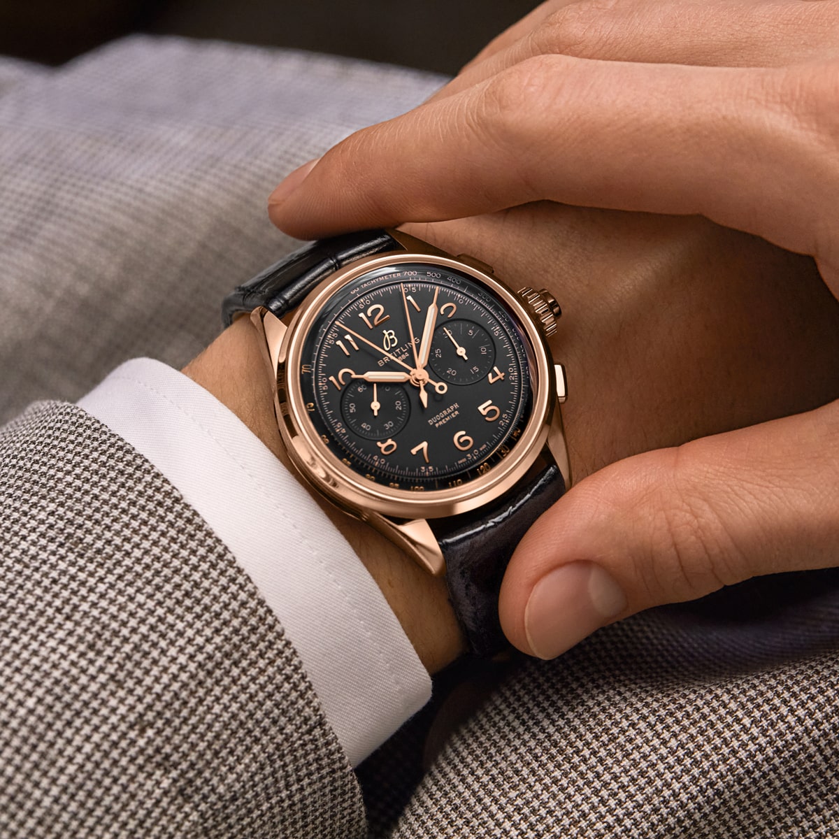 2025 © BREITLING Nahaufnahme einer Person, die eine goldene Breitling-Armbanduhr mit schwarzem Lederarmband und detailliertem schwarzem Zifferblatt trägt, bekleidet mit einer braun karierten Anzugsjacke und einem weißen Hemd. Die Hand stellt die Uhr ein.