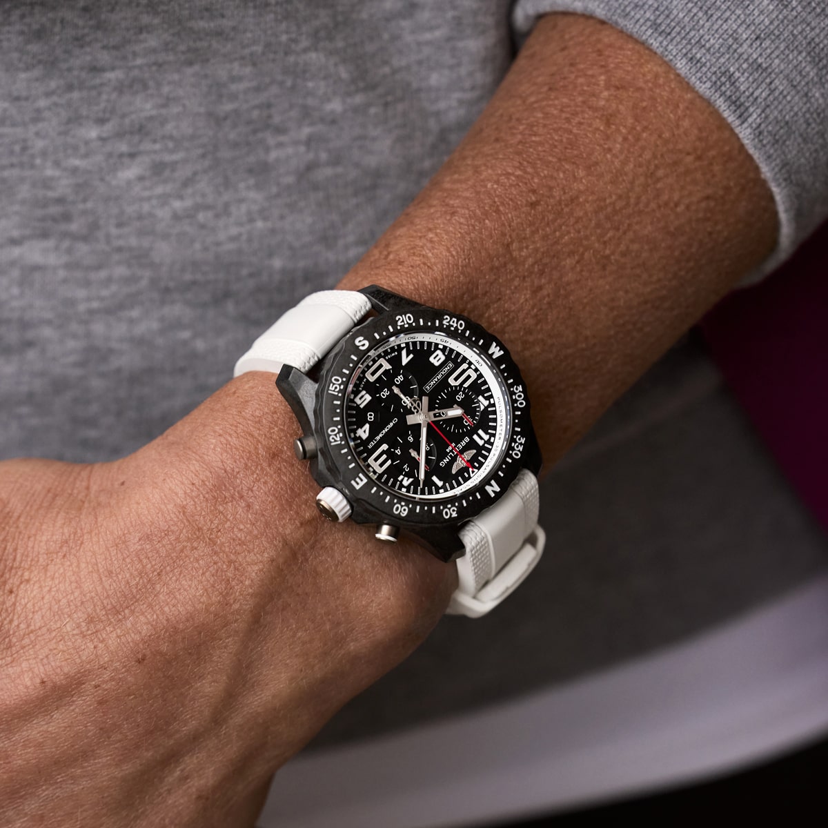 2025 © BREITLING Eine Person trägt eine schwarze Breitling-Uhr mit einem weißen Band am Handgelenk. Das Zifferblatt weist weiße Markierungen, mehrere Ziffernblätter und Kompassrichtungen auf der Lünette auf. Die Person ist mit einem grau-weißen Oberteil bekleidet.