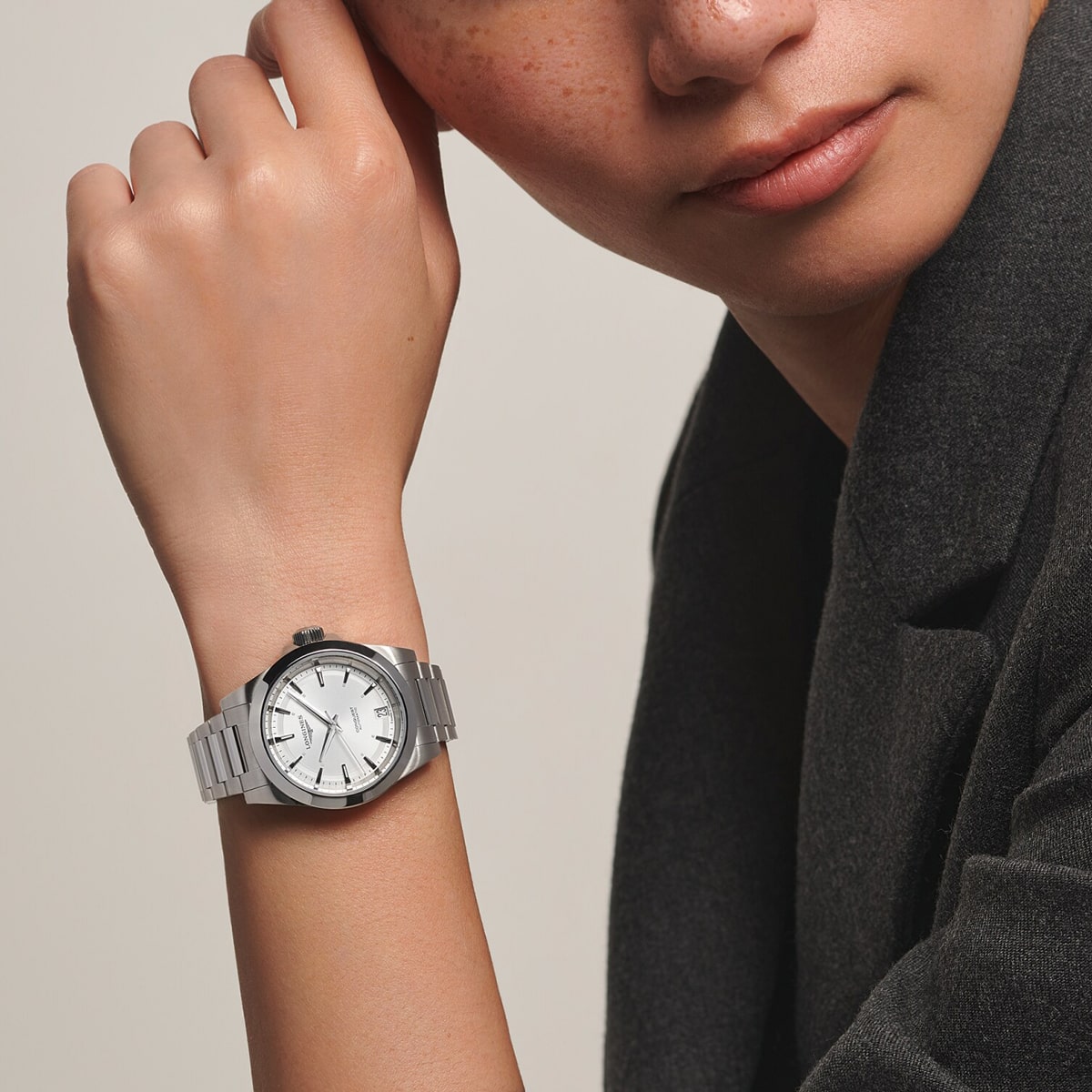 2025 © LONGINES Eine Person, die einen dunklen Blazer trägt, stützt ihr Kinn auf die Hand und zeigt eine silberne Conquest-Armbanduhr mit weißem Ziffernblatt und Metallband, die sie am linken Handgelenk trägt.