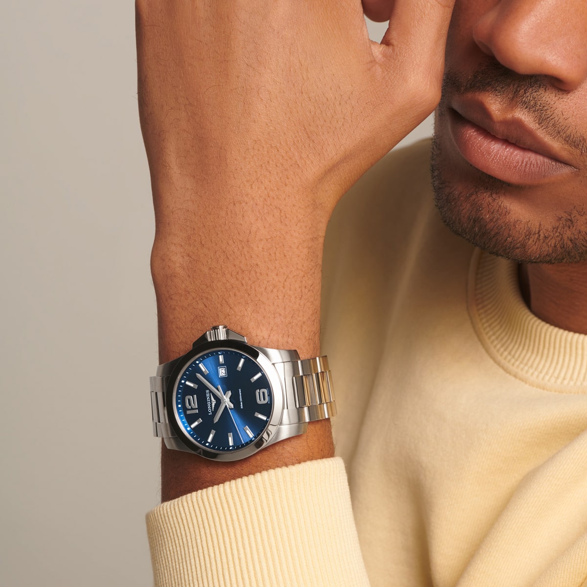 2025 © LONGINES Eine Person, die einen hellgelben Pullover trägt, zeigt eine silberne Conquest-Armbanduhr mit blauem Zifferblatt und Metallband an ihrem linken Handgelenk. Die Uhr ist gut zu erkennen, während das Gesicht der Person nur teilweise sichtbar ist.