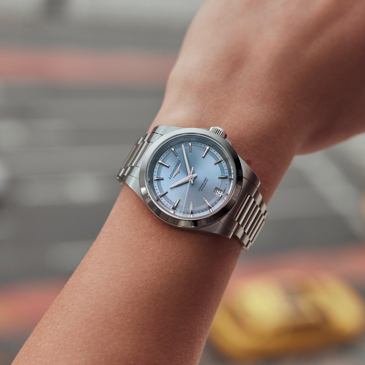 2025 © LONGINES Nahaufnahme eines Handgelenks, das eine silberne Conquest-Uhr mit blauem Zifferblatt und silbernen Stundenmarkierungen trägt, fotografiert im Freien über einer verschwommenen Straße mit gelben Autos im Hintergrund.