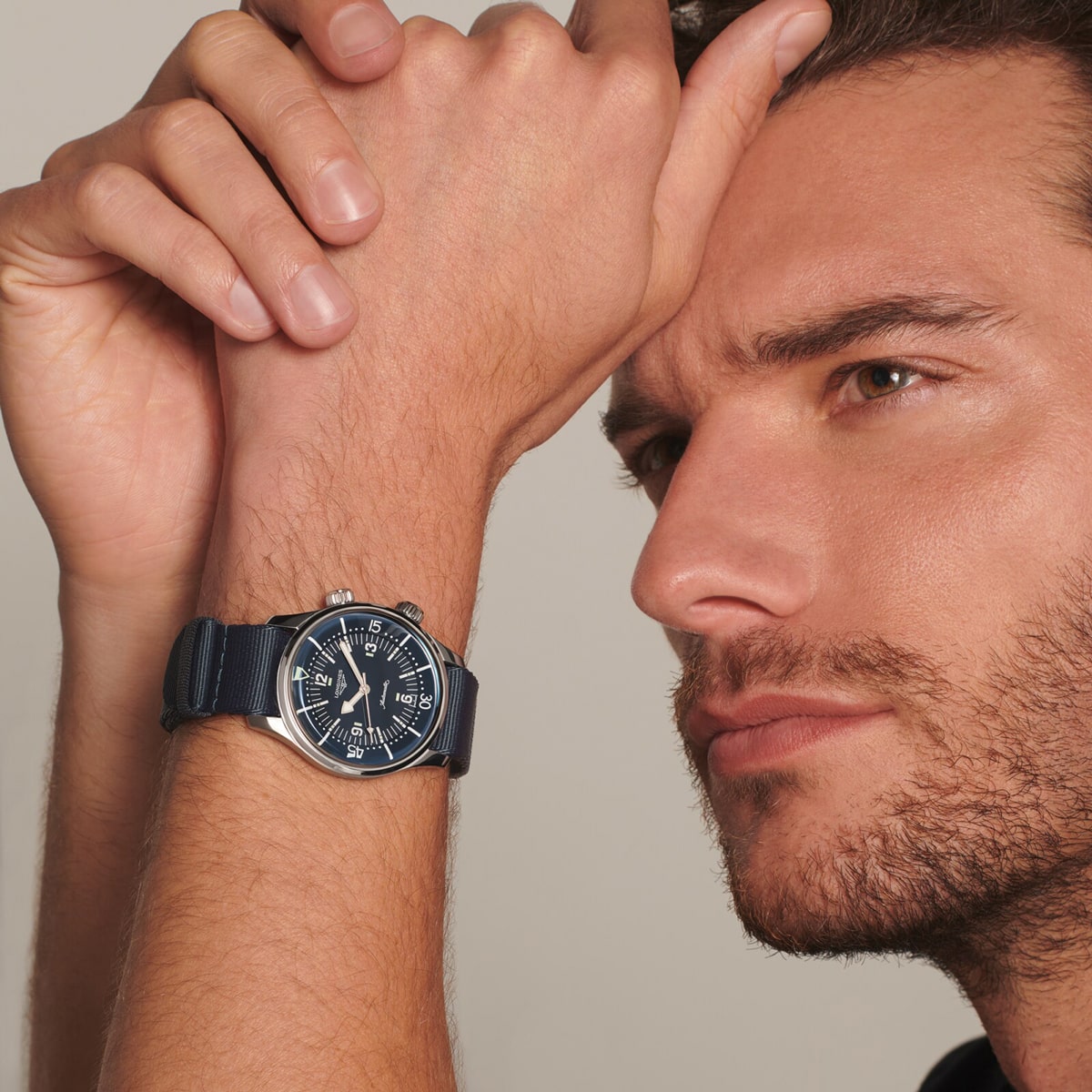2025 © LONGINES Ein Mann mit kurzem braunem Haar und gestutztem Bart hält seinen Arm hoch und zeigt eine dunkelblaue Legend Diver-Armbanduhr mit rundem Zifferblatt und silbernen Details. Vor einem neutralen Hintergrund blickt er nachdenklich in die Ferne.