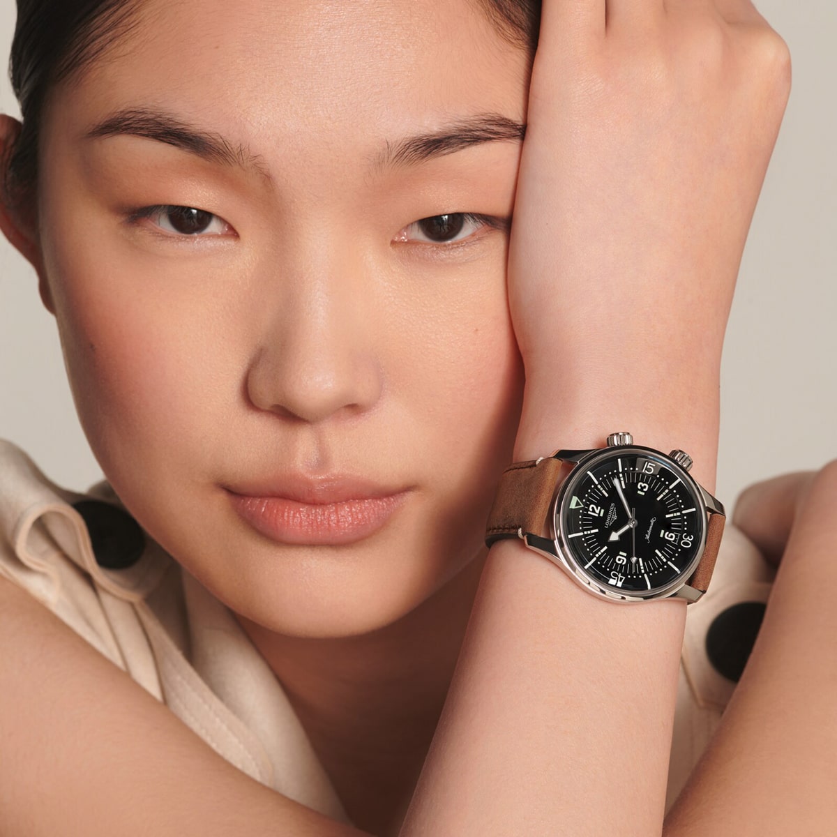 2025 © LONGINES Eine Frau mit glattem schwarzem Haar stützt ihr Gesicht auf ihren Unterarm und zeigt eine silberne Legend Diver-Uhr mit schwarzem Zifferblatt und hellbraunem Lederarmband. Sie blickt ruhig in die Kamera vor einem neutralen Hintergrund.