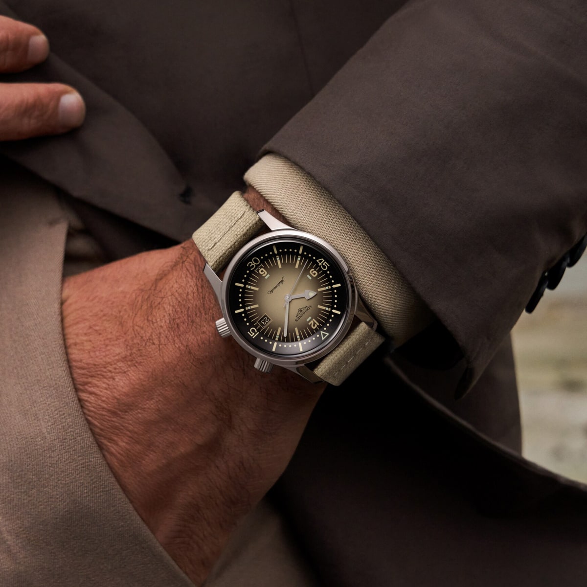 2025 © LONGINES Eine Person, die einen braunen Mantel und eine beigefarbene Manschette trägt, zeigt eine Legend Diver Armbanduhr mit einem beigen Armband und einem runden, dunklen Zifferblatt mit auffälligen weißen Stundenmarkierungen und Zeigern. Die Uhr hat ein klassisches, elegantes Design.