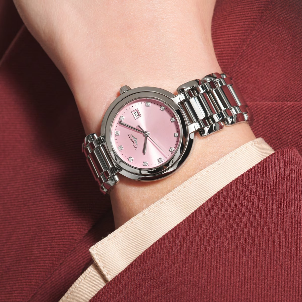2025 © LONGINES Nahaufnahme eines Handgelenks mit einer silbernen PrimaLuna-Uhr mit rosafarbenem Ziffernblatt und Diamantindexen, kombiniert mit einer burgunderroten Anzugsjacke und einem cremefarbenen Hemd.