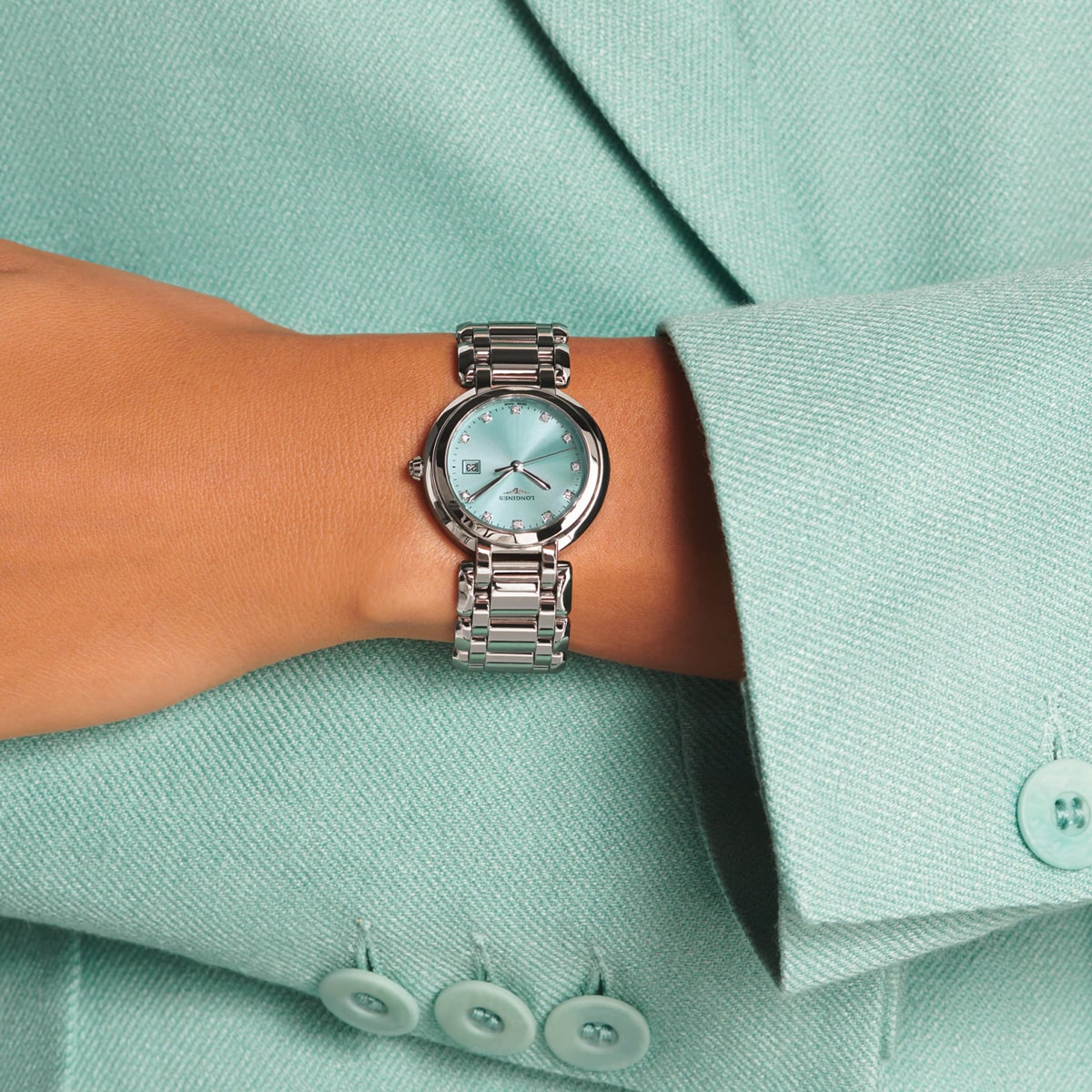 2025 © LONGINES Eine Person, die eine hellblaue Jacke mit drei Knöpfen trägt, zeigt eine silberne PrimaLuna-Armbanduhr mit einem passenden hellblauen Zifferblatt an ihrem linken Handgelenk.