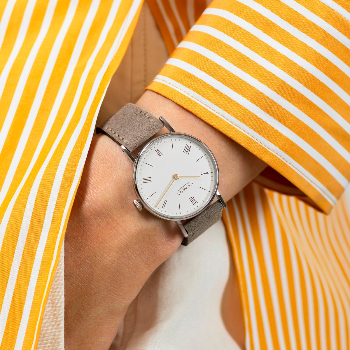 2025 © NOMOS GLASHÜTTE Eine Person, die eine NOMOS Glashütte Ludwig mit grauem Lederarmband und weißem Zifferblatt mit römischen Ziffern trägt, bekleidet mit einem gelb-weiß gestreiften langärmeligen Hemd.