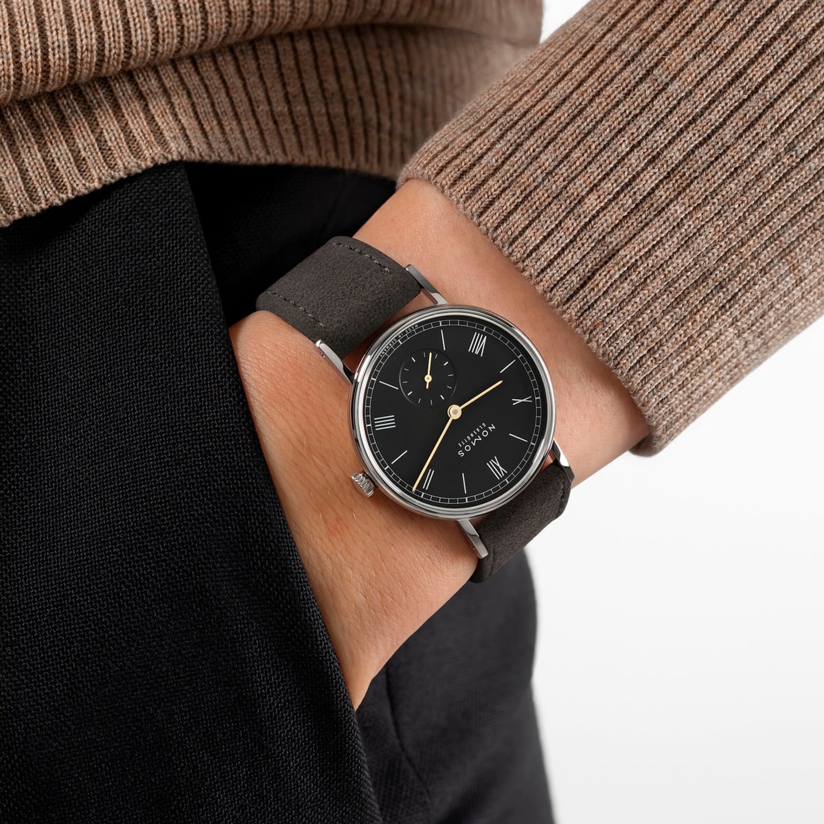 2025 © NOMOS GLASHÜTTE Eine Person, die einen braunen Rippenpullover und eine dunkle Hose trägt, hat die linke Hand in der Tasche und zeigt eine NOMOS Glashütte Ludwig-Armbanduhr mit römischen Ziffern und schwarzem Lederband.