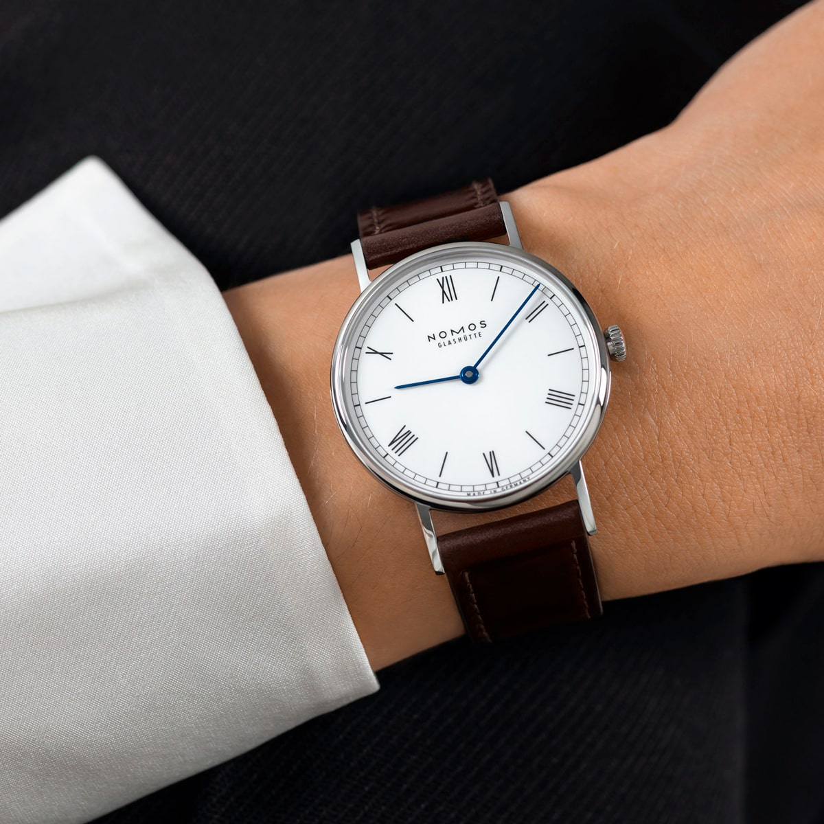 2025 © NOMOS GLASHÜTTE Eine Person, die ein weißes Hemd mit Manschette und eine dunkle Jacke trägt, zeigt eine NOMOS Glashütte Ludwig-Armbanduhr mit weißem Zifferblatt, römischen Ziffern, blauen Zeigern und braunem Lederarmband.