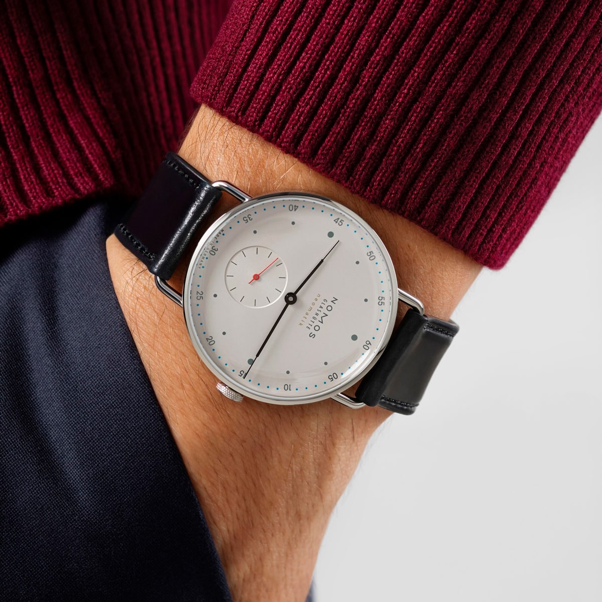 2025 © NOMOS GLASHÜTTE Eine Person trägt eine minimalistische silberne NOMOS Glashütte Metro-Armbanduhr mit weißem Zifferblatt, kleiner Sekundenanzeige und schwarzem Lederarmband. Ihre Hand steckt in der Tasche, und sie ist mit einem burgunderroten Pullover bekleidet.