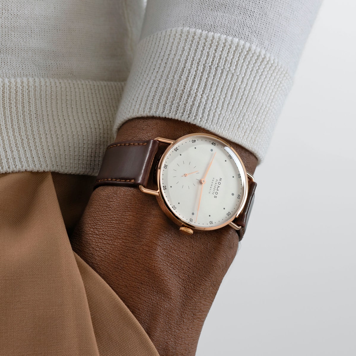 2025 © NOMOS GLASHÜTTE Eine Nahaufnahme des Handgelenks einer Person, die eine stilvolle NOMOS Glashütte Metro-Uhr mit weißem Zifferblatt und minimalistischem Design trägt. Die Person ist mit einem hellen Pullover und einer hellbraunen Hose bekleidet.