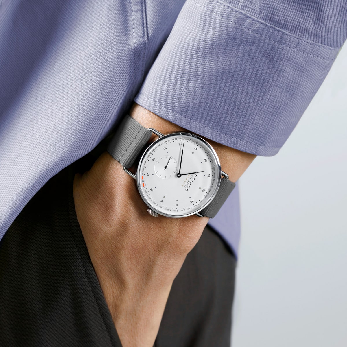 2025 © NOMOS GLASHÜTTE Eine Person, die ein hellblaues langärmeliges Hemd und eine dunkle Hose trägt, hat eine minimalistische NOMOS Glashütte Metro-Armbanduhr mit weißem Zifferblatt und grauem Armband am linken Handgelenk; die Hand steckt in der Tasche.