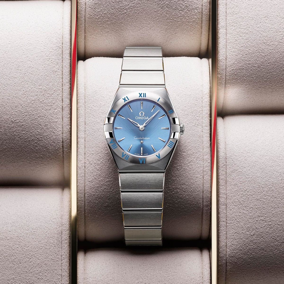 2025 © OMEGA Eine Omega Constellation Armbanduhr aus Edelstahl mit blauem Zifferblatt und römischen Ziffern befindet sich in einem gepolsterten Uhrengehäuse. Das Metallarmband ergänzt das Zifferblatt, das das Omega-Logo und den Text "Constellation" zeigt.