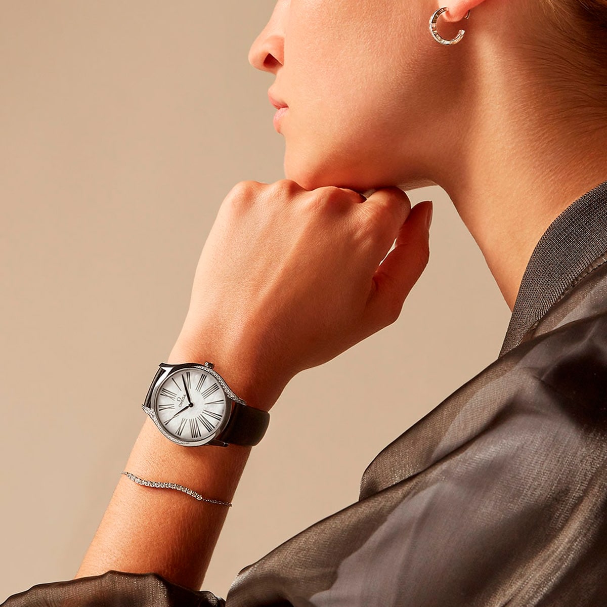 2025 © OMEGA Eine Person, die eine De Ville-Armbanduhr mit schwarzem Lederarmband und rundem Silberzifferblatt, einen Ohrring und ein zartes Armband trägt, bekleidet mit einem durchsichtigen dunklen Oberteil, posiert mit der Hand in der Nähe ihres Gesichts vor einem beigen Hintergrund.