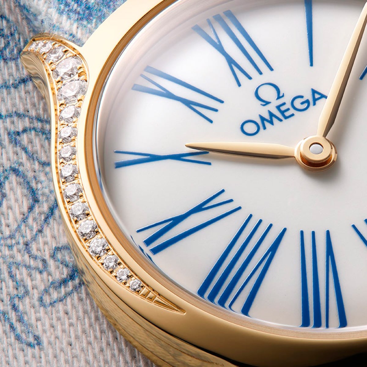 2025 © OMEGA Nahaufnahme einer Omega De Ville-Armbanduhr mit einem goldenen Gehäuse, blauen römischen Ziffern auf einem weißen Zifferblatt, goldenen Zeigern und einer Reihe kleiner Diamanten entlang der Lünette. Im Hintergrund ist ein Teil eines strukturierten Stoffes zu sehen.