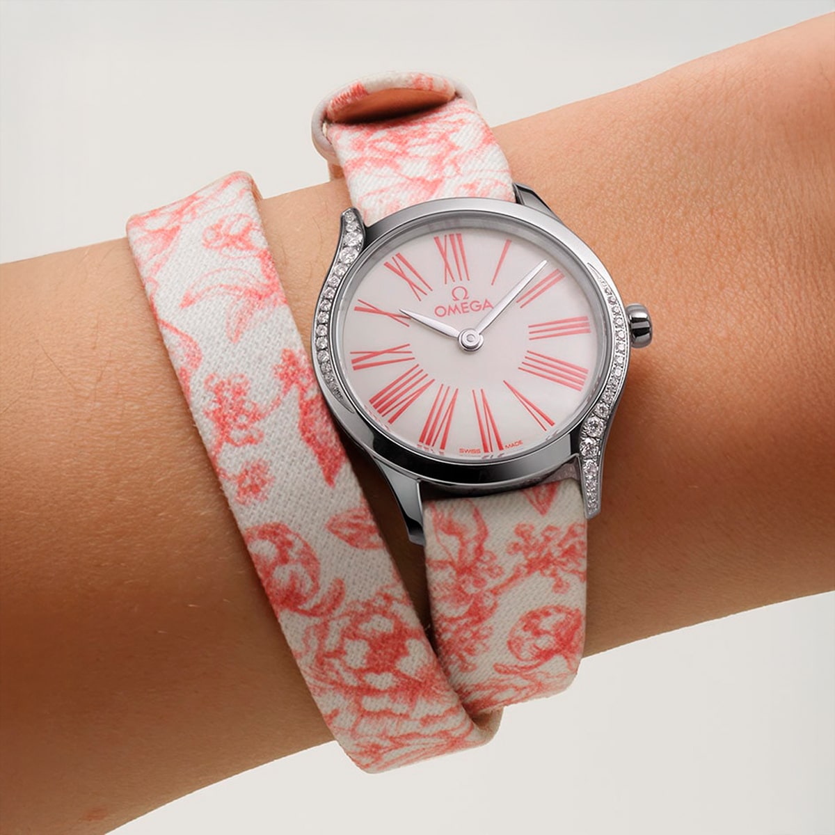 2025 © OMEGA Ein Handgelenk trägt eine Omega De Ville-Uhr mit silbernem Gehäuse, weißem Zifferblatt, rosafarbenen römischen Ziffern und einem rosafarbenen, mit Blumen gemusterten Stoffarmband.