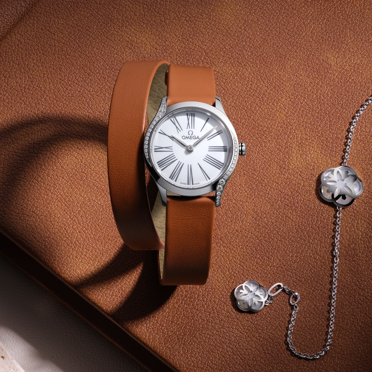 2025 © OMEGA Eine silberne Omega De Ville-Armbanduhr mit weißem Zifferblatt und braunem Lederarmband wird auf einer strukturierten braunen Oberfläche neben einer silbernen kleeblattförmigen Halskette und einem passenden Anhänger präsentiert.