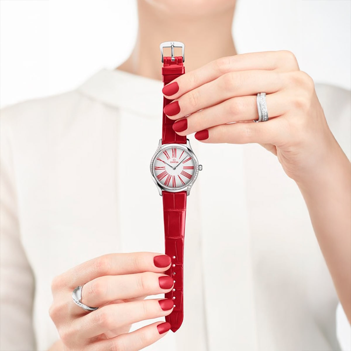 2025 © OMEGA Eine Frau mit rot lackierten Nägeln und silbernen Ringen hält eine De-Ville-Armbanduhr mit rotem Lederarmband und rot-silbernem Zifferblatt in der Hand und trägt eine weiße Bluse.